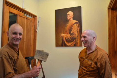 Two monks working on fixing a door with a portrait of Ajahn Sucitto on the wall.