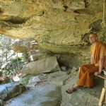 Ajahn Medhino sits under the broad cave-like overhang. Warm and dry.