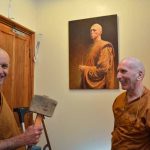Two monks working on fixing a door with a portrait of Ajahn Sucitto on the wall.